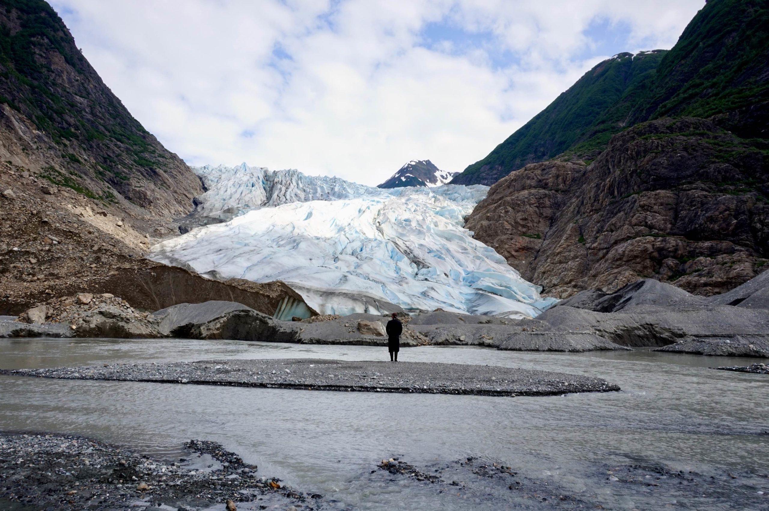 A person standing on a small island in a stream in front of a glacier