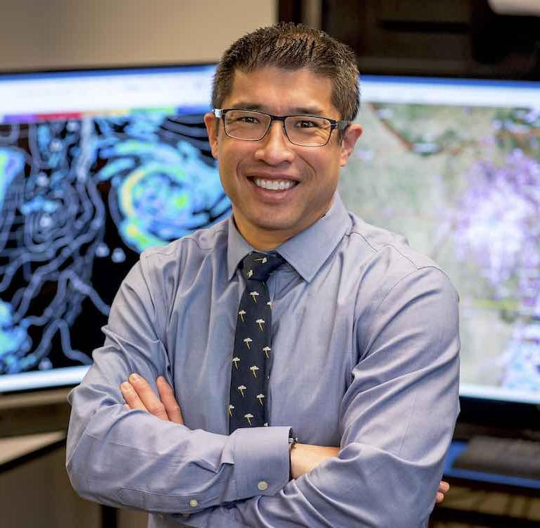 Dr. Sam Ng standing in front of two computer monitors displaying weather maps