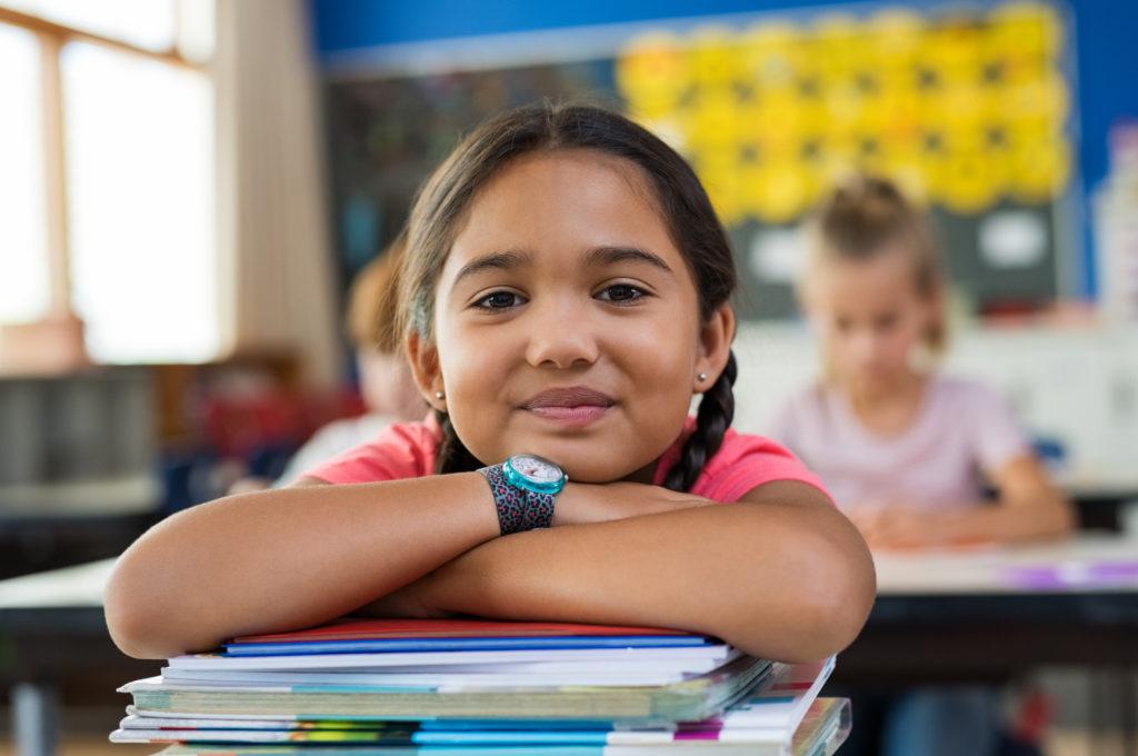 Portrait of little schoolgirl leaning on stacked books in classroom.