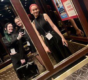 Three students photograph their reflections in a mirror, they are wearing conference badges.