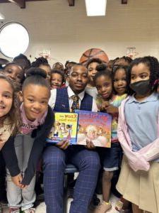 A photo of one of the MSU Denver MISTERs sitting with a group of young students and holding two books.