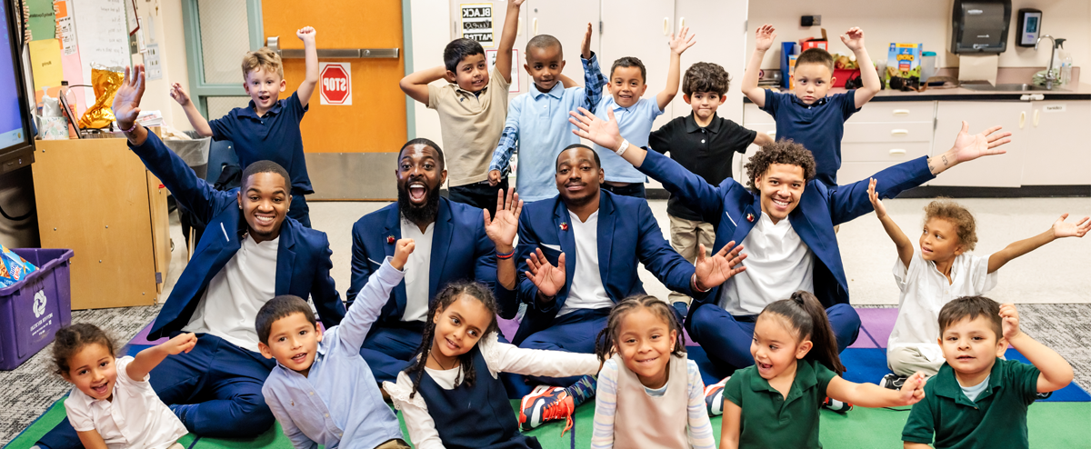 The MSU Denver Call Me MISTER students and director sit with a group of elementary school students, arms extended and smiling.