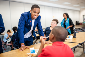 Call Me MISTER student Josh Barringer stands smiling across the lunch table from a young student in an elementary school cafeteria.