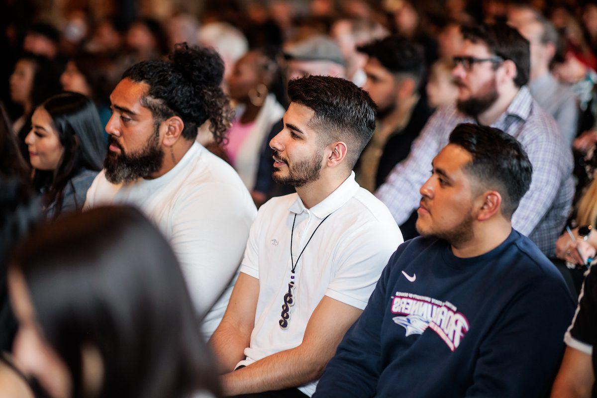 three male students of color sitting in a row of seats at an event slightly smiling