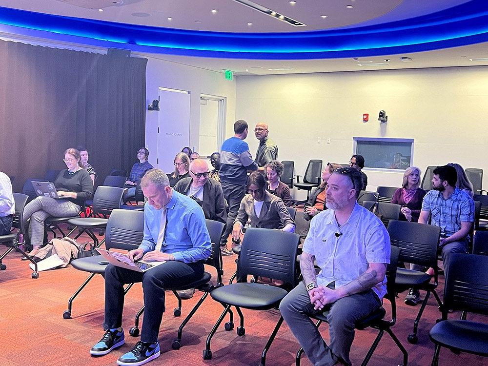 The image shows the audience at Travis Masingale's presentation on using AI in college classrooms, an event sponsored by the Department of English at Metropolitan State University Denver and held in the CAVEA. The attendees, who are primarily faculty members, are seated in rows of chairs, some taking notes on laptops or notebooks. The room has a curved layout with blue LED lighting along the edges of the ceiling.