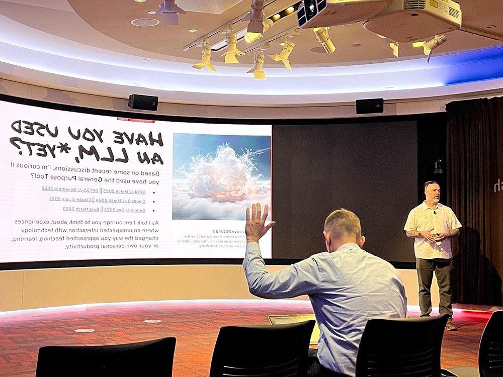 The image shows Travis Masingale from Eastern Washington University giving his presentation on using AI in college classrooms to faculty members. The event is sponsored by the Department of English at Metropolitan State University Denver and is taking place in the CAVEA. Mr. Masingale is standing at the front of the lecture hall, gesturing toward his slide projected on the screen behind him. The slide asks 