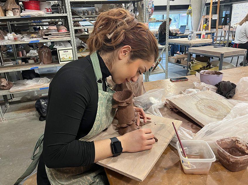 Female student working on an unfired hand built vessel in the ceramics classroom with view of storage shelves in the background .