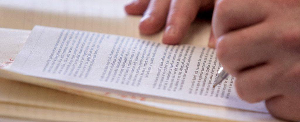 A close-up of a person's hand writing on a small piece of paper with printed text, resting on a larger lined notepad.