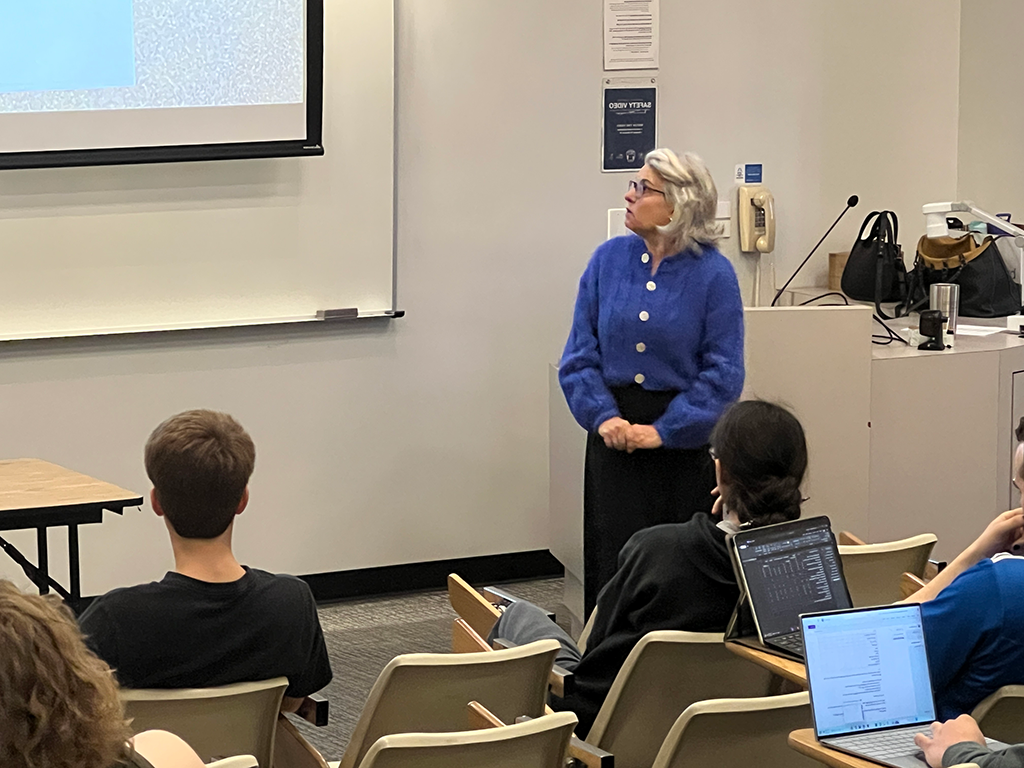 Professor Pamela Troyer, wearing a blue sweater, stands at the front of a classroom giving a lecture. Several students are seated facing her, some with laptops open. The classroom has a projector screen and a lectern with various items on it.