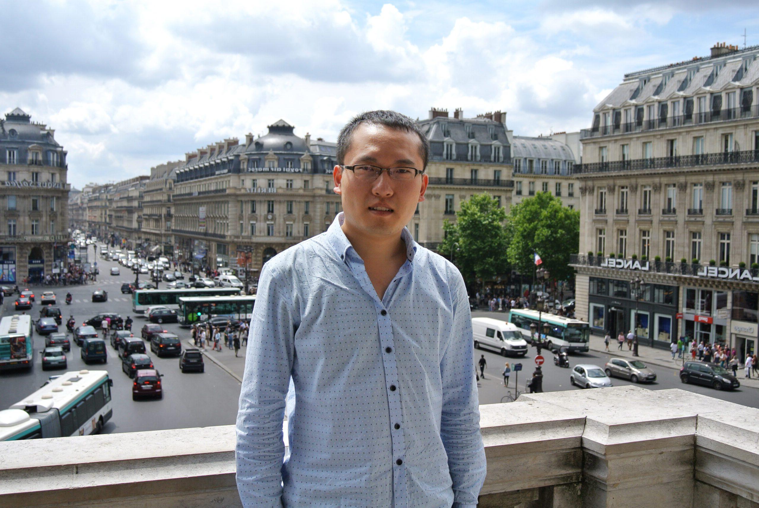 Dr. Andong Ma standing on a balcony with a european city center behind him