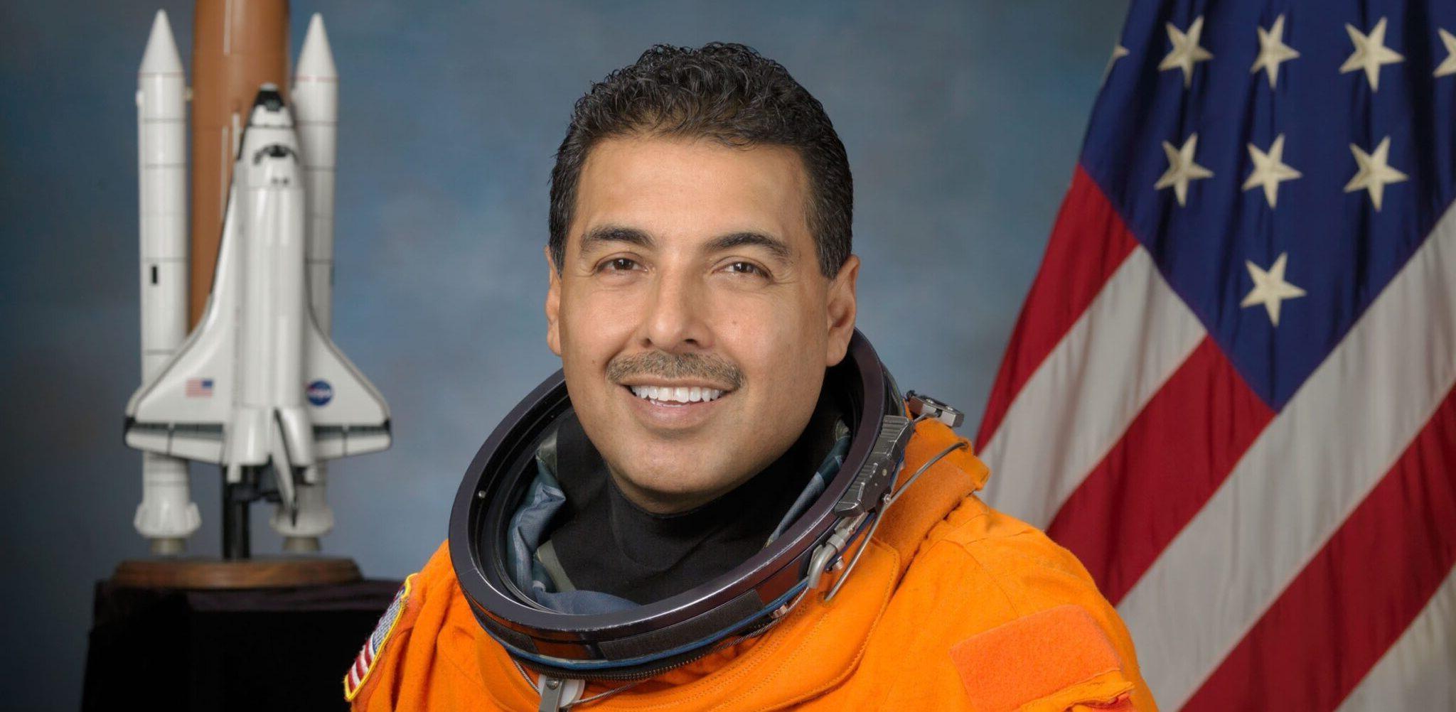 Astronaut Jose M. Hernandez, 身穿橙色太空服的任务专家, 背景是美国国旗和航天飞机模型