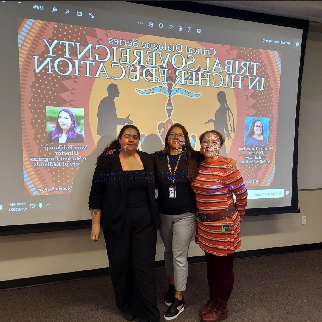 Three Native American staff members pose in front of an event flyer. Event Flyer text: Tribal Sovereignty in Higher Education